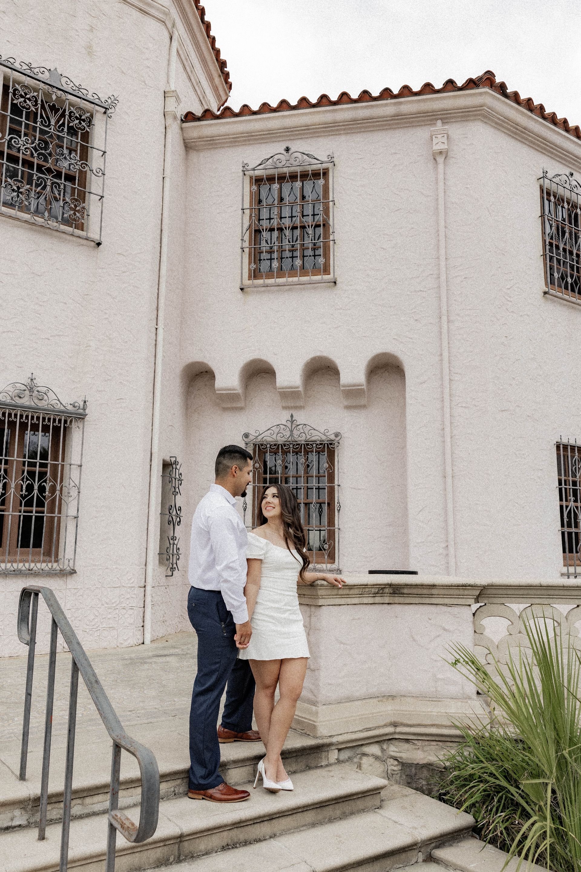 Couple standing on steps outside a white stucco house with barred windows and red-tiled roof