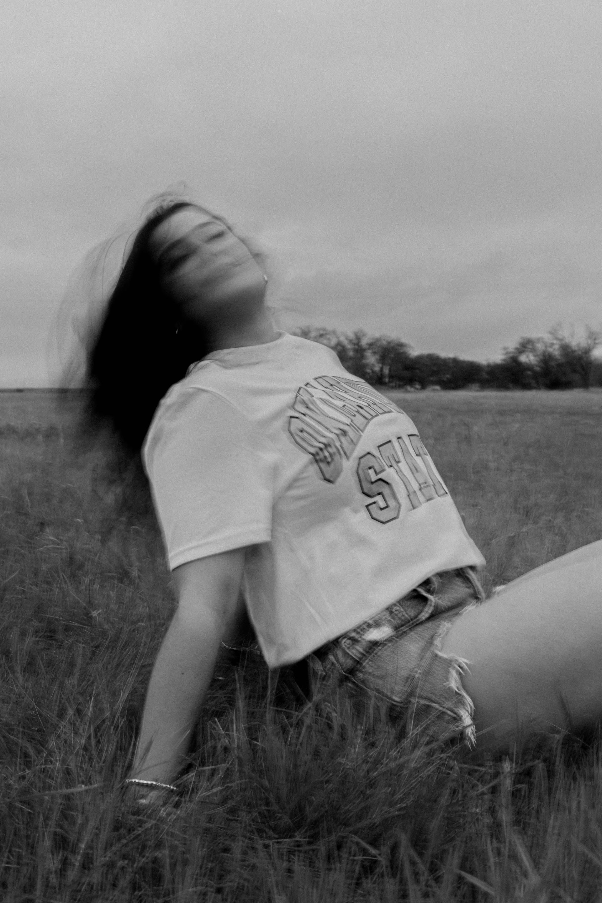 Person reclining in tall grass, leaning back with head tilted up in a black-and-white outdoor scene