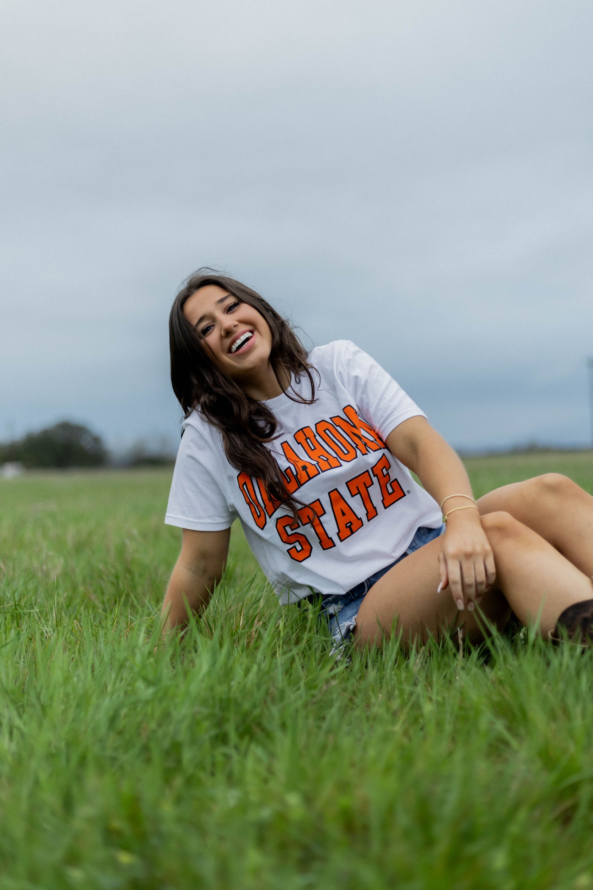 Woman in a white “Texas State” T-shirt sitting in green grass, smiling under a cloudy sky.