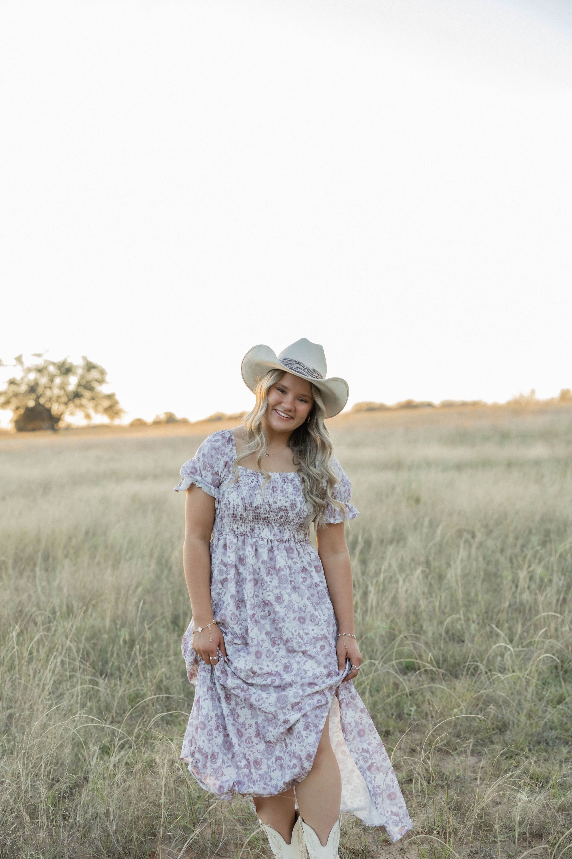 Woman in a floral dress and cowboy hat standing in a grassy field at sunset
