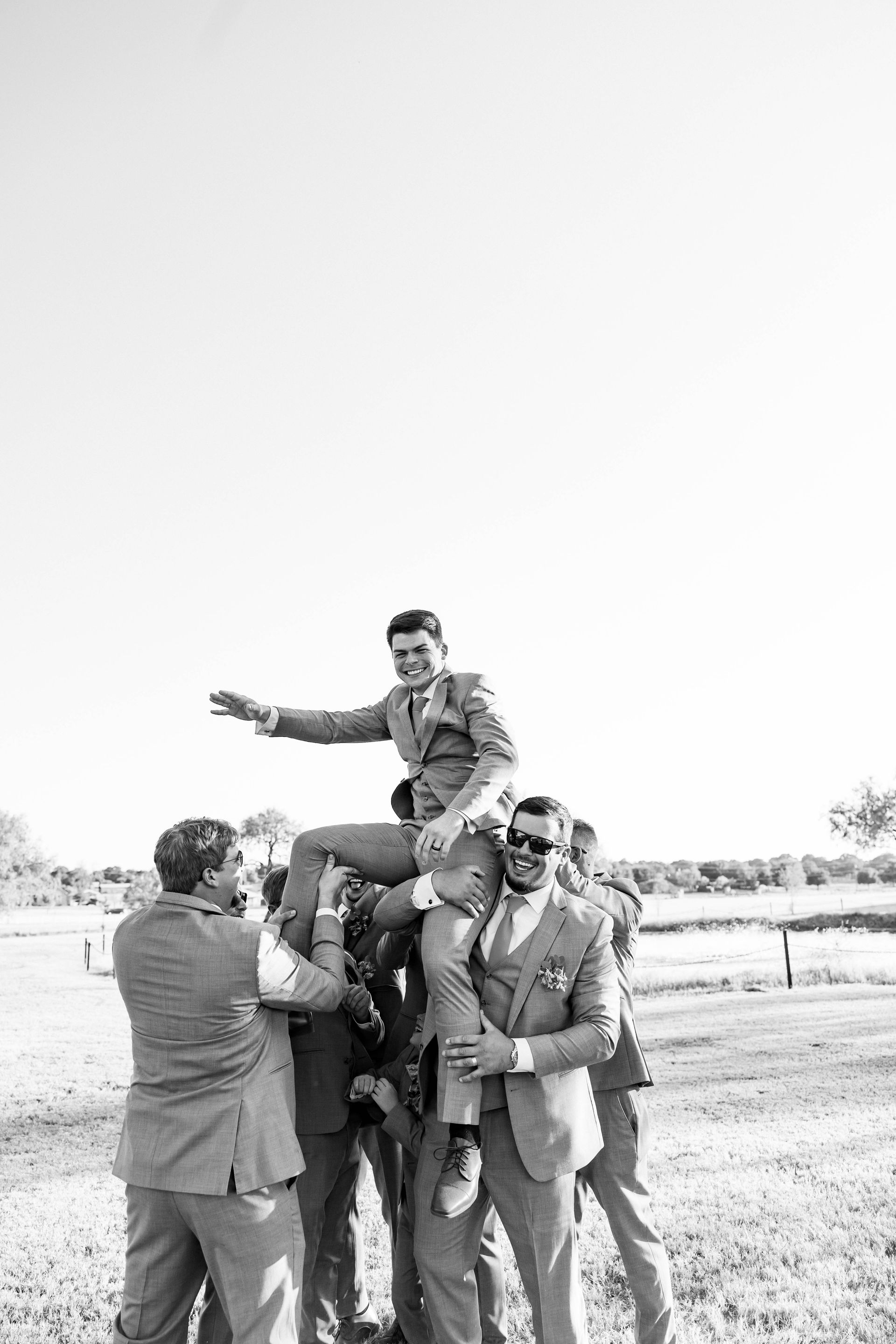 Group of men in suits lifting another man on their shoulders outdoors, with one pointing ahead.