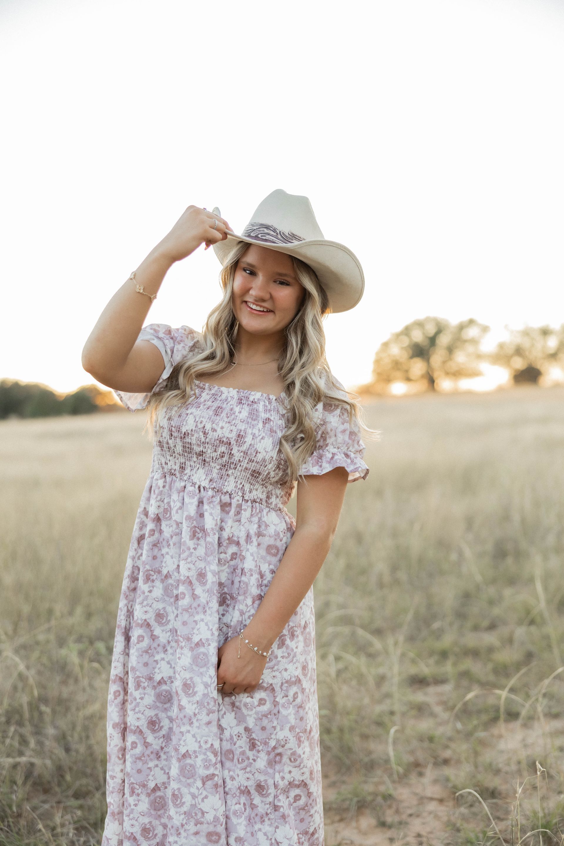 Woman in a white floral dress and cowboy hat standing in a grassy field, smiling at sunset