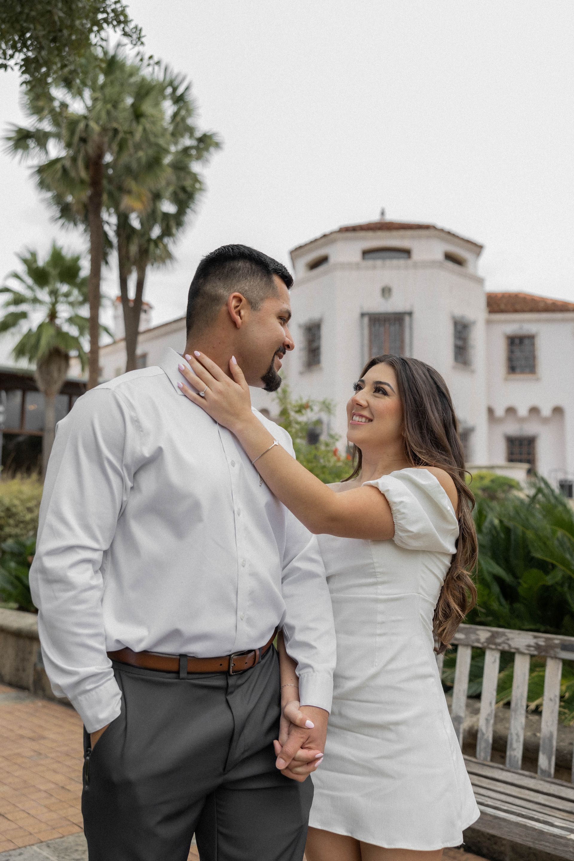 Couple holding hands and smiling outdoors near a white building and palm trees