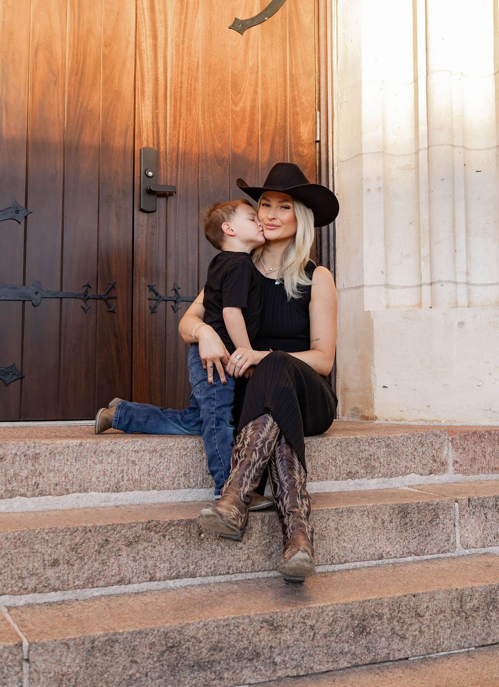Person and child sitting on stone steps by a wooden door, wearing black clothes and a hat