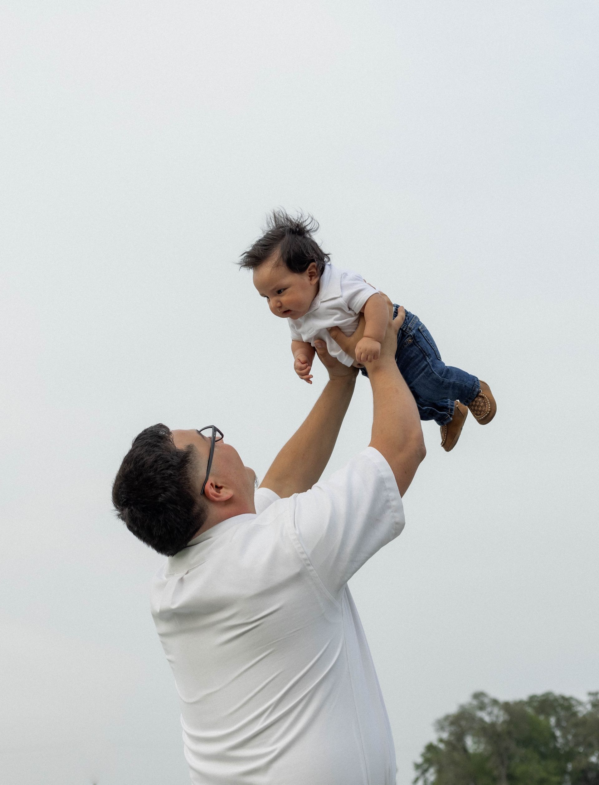 Man in white shirt lifting a small child into the air outdoors against a cloudy sky