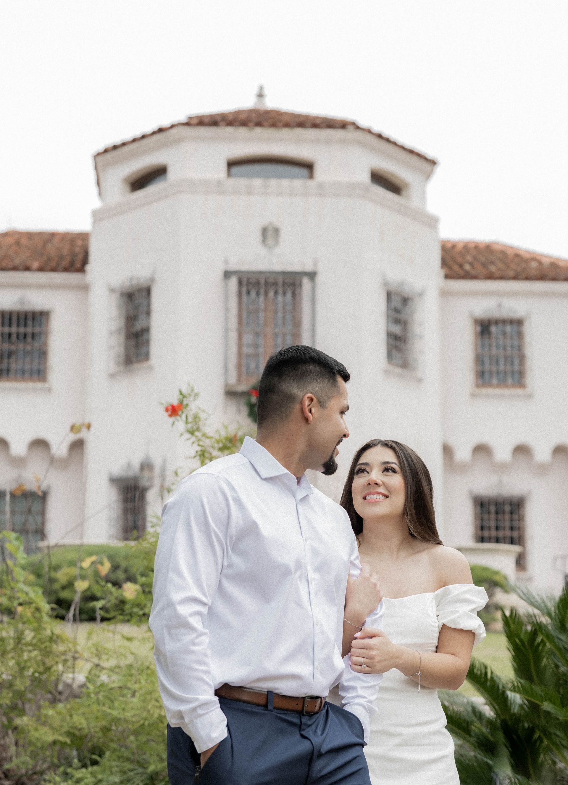 Couple in white clothing standing outside a white stucco building, smiling at each other.