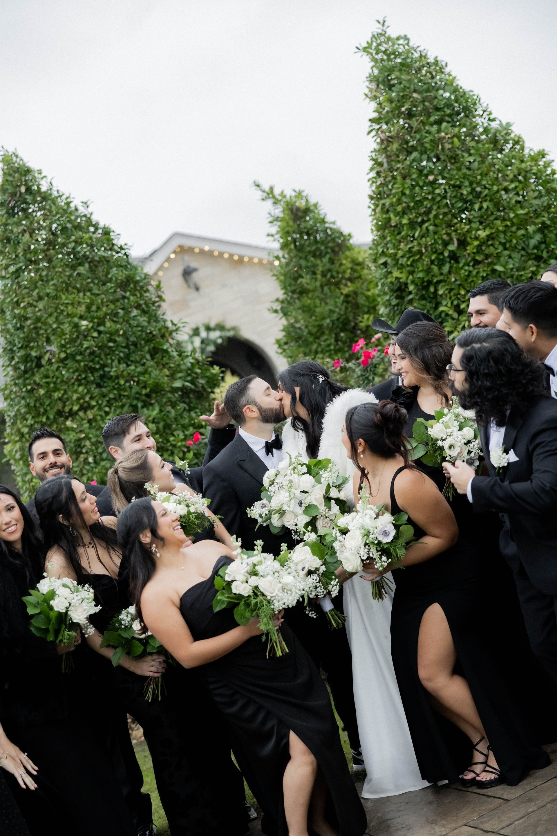 Wedding party in black attire surrounds a bride in white outdoors by a stone archway and tall hedges