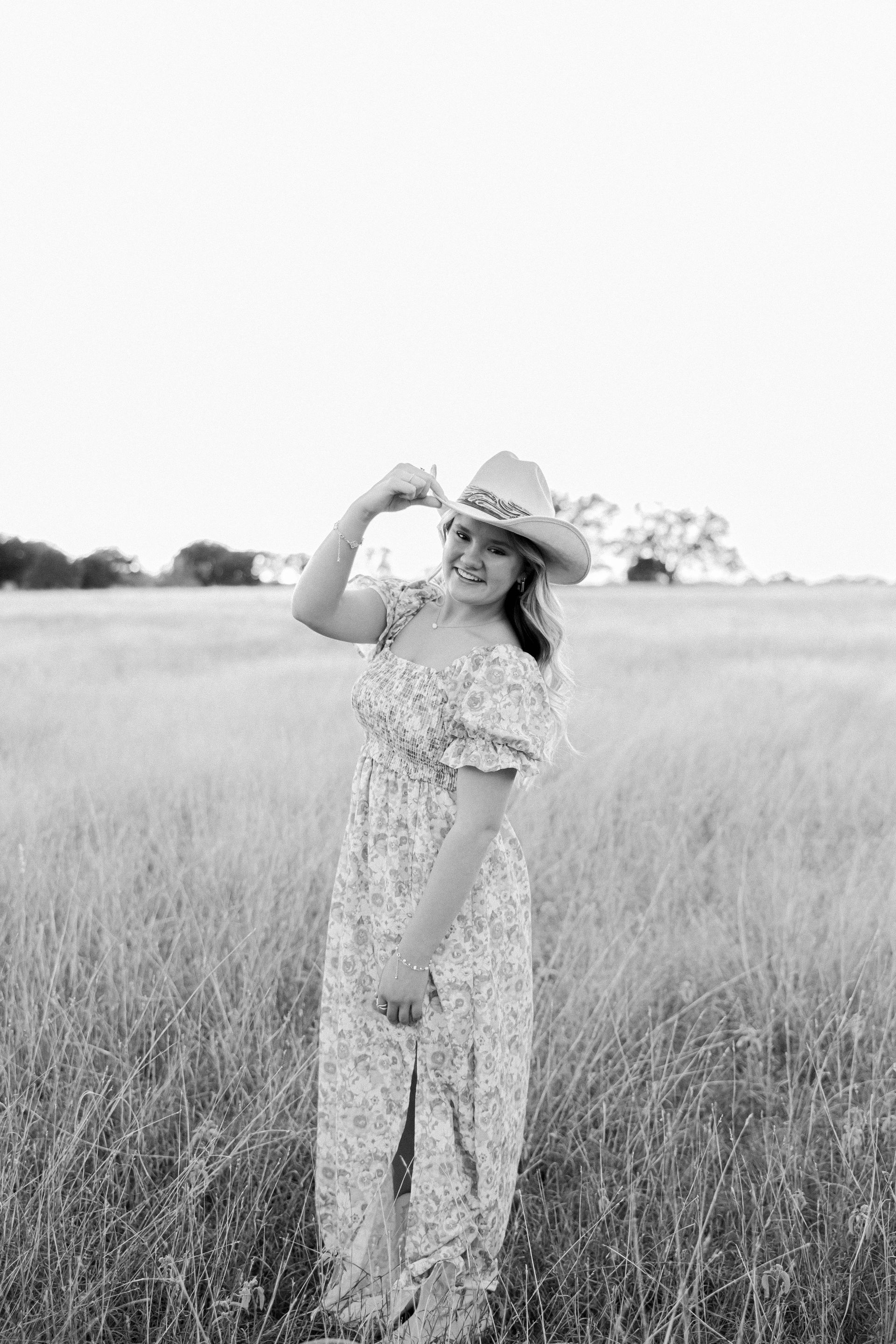 Person in a floral jumpsuit and hat standing in a grassy field, posing with one hand raised.