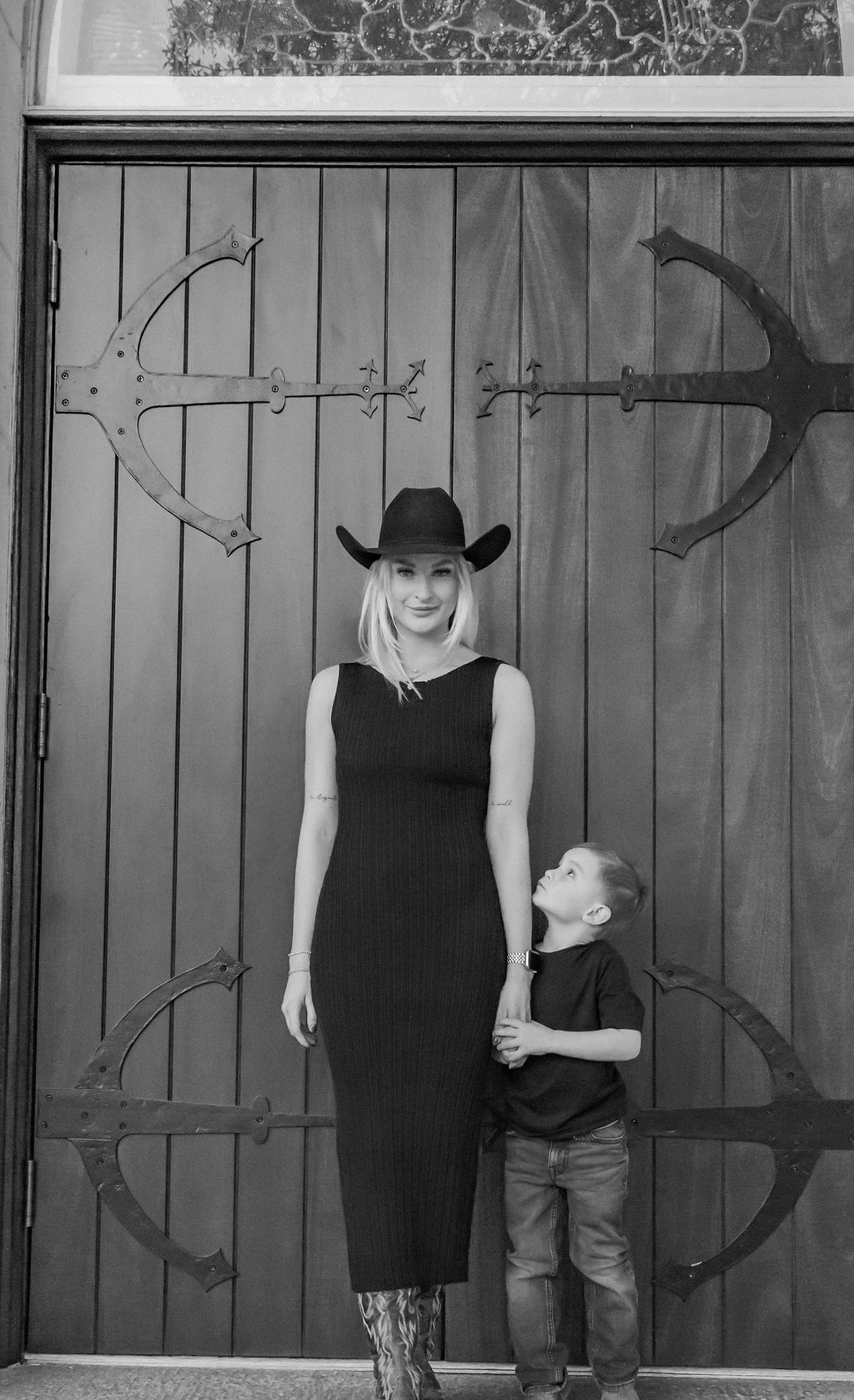 Woman in a black dress and child standing in front of a wooden gate, both wearing hats.