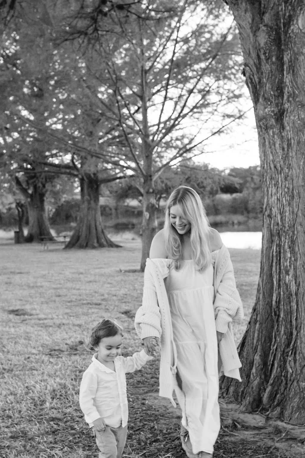 Woman and child walking hand in hand beside a tree in a park, both smiling.