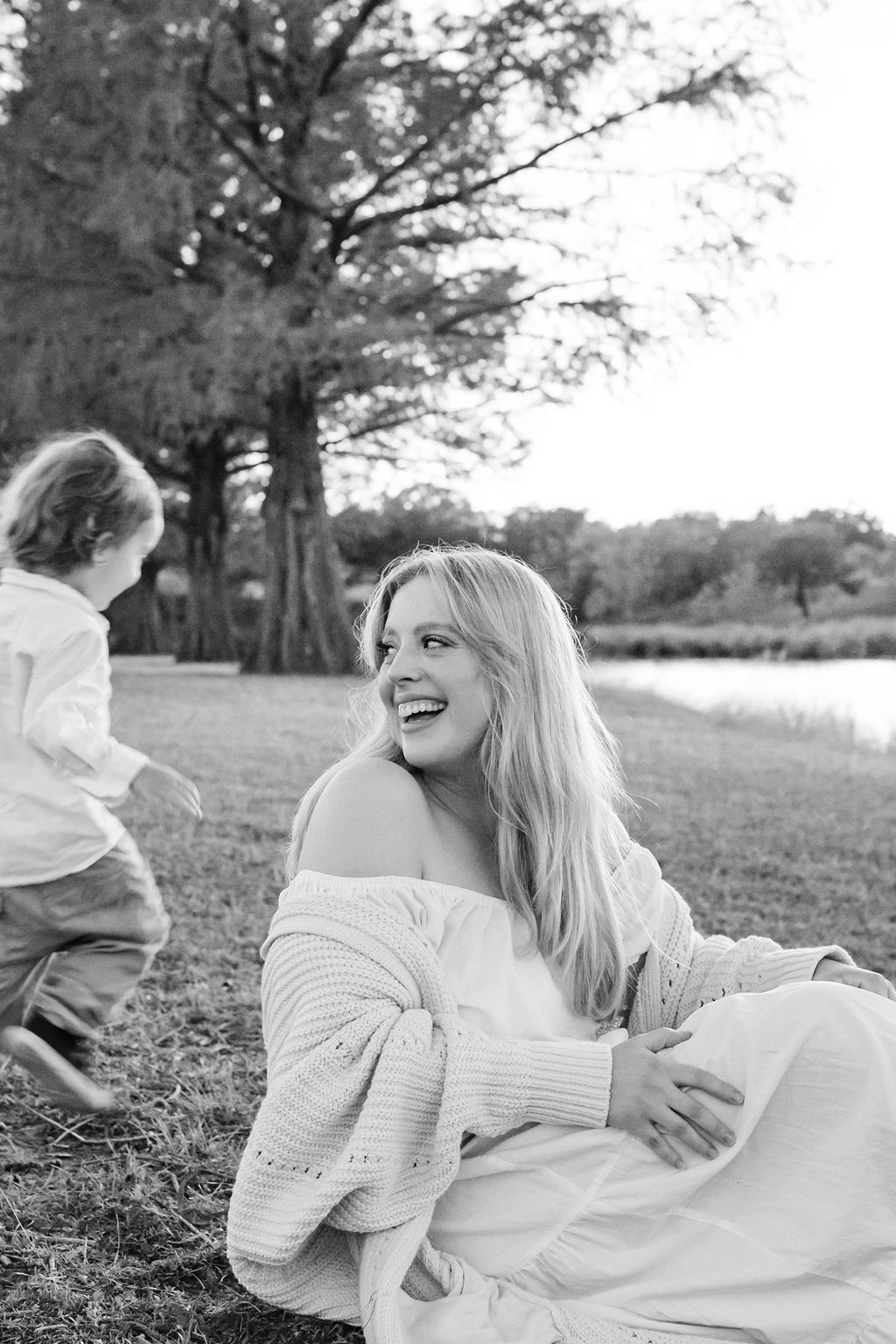 Woman sitting on grass by a lakeside, smiling at a child walking nearby under trees.
