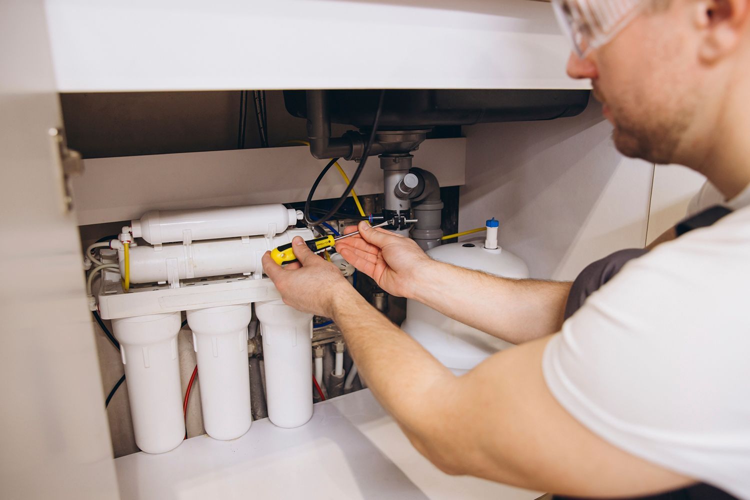 A plumber installing a water softening system under the kitchen sink. A plumber installing a water softening system under the kitchen sink.