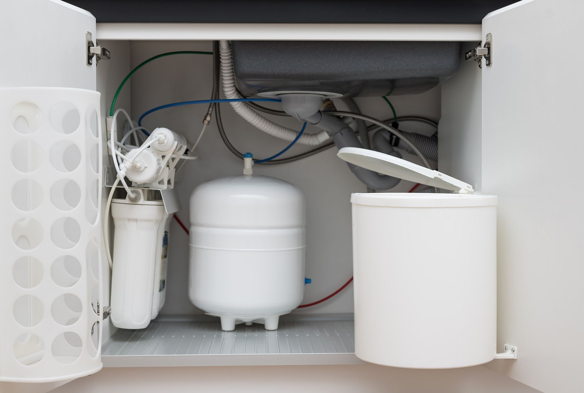 Under-sink water filtration system with storage tank, filters, and connected tubing inside a kitchen cabinet. Under-sink water filtration system with storage tank, filters, and connected tubing inside a kitchen cabinet.