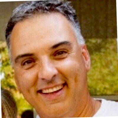 Man smiling, looking towards the camera. He has short gray hair and fair skin.