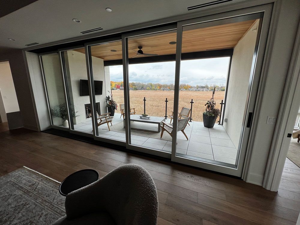 Sliding glass doors open to a patio with seating, fireplace, and a distant field. Interior has wood floors and a chair.
