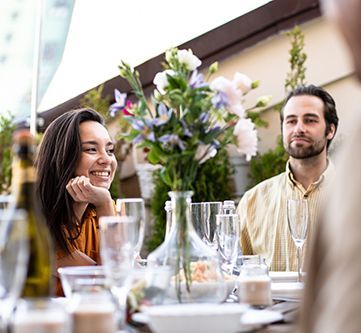 People seated at an outdoor table, smiling and looking at each other, with flowers and champagne glasses.