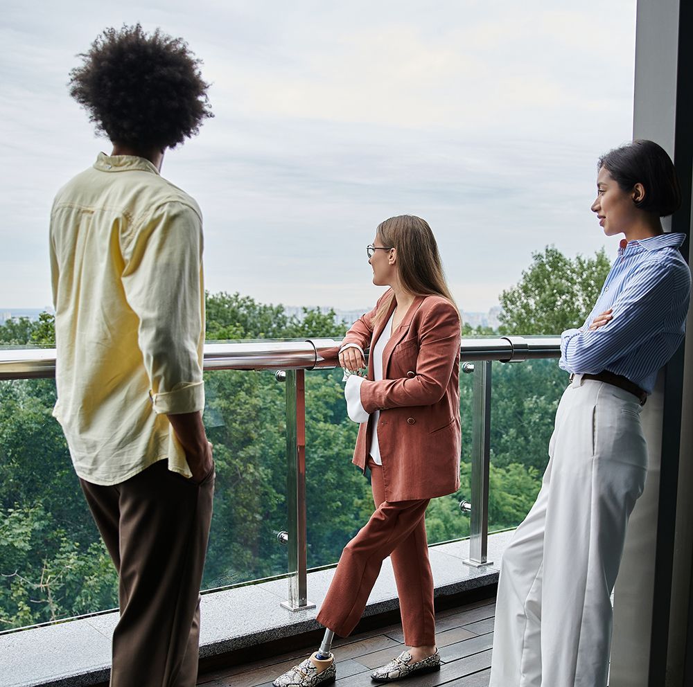 Three people on a balcony overlooking trees and cloudy sky. One has a prosthetic leg, all leaning on railing.