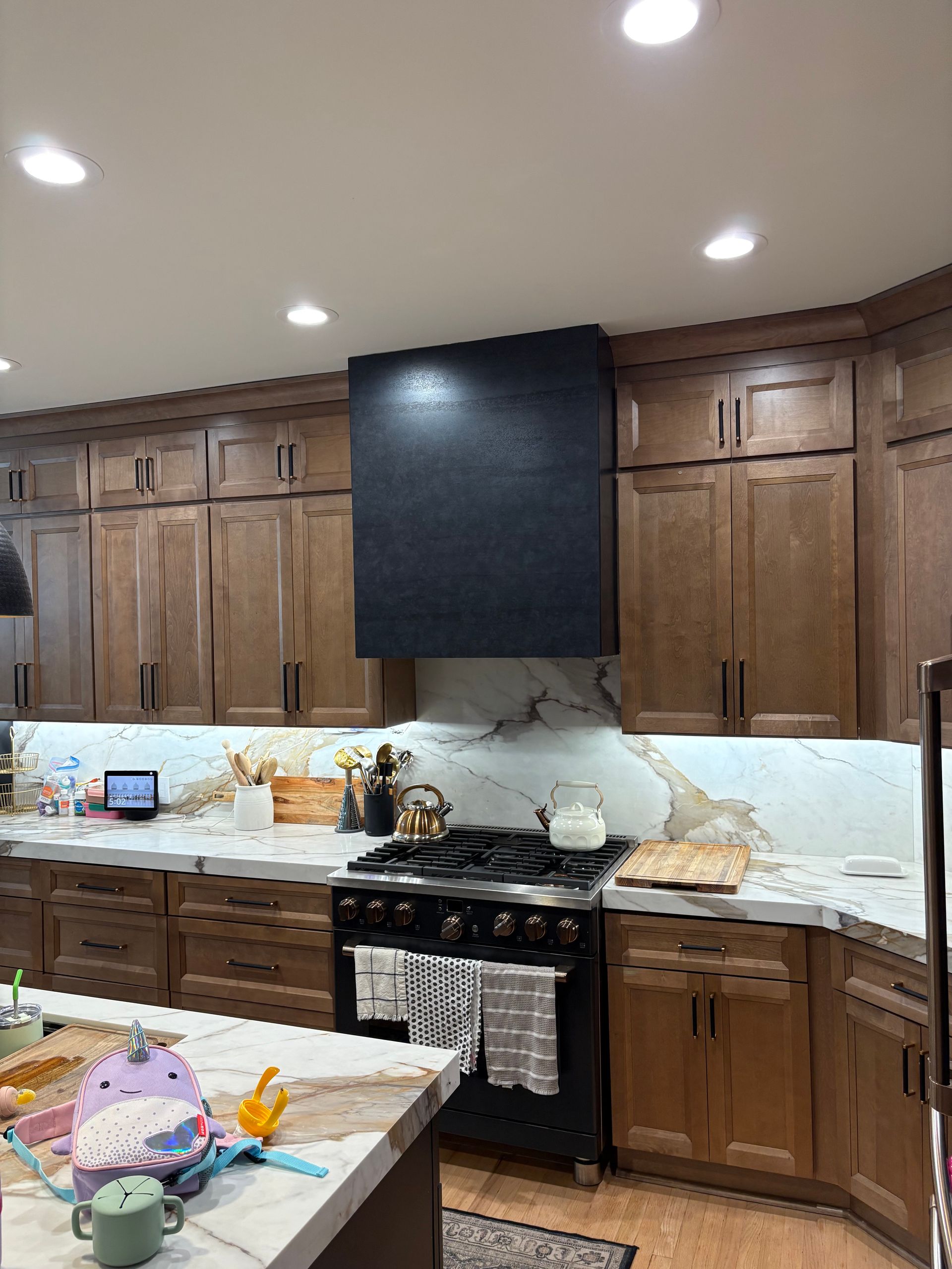 Modern kitchen with dark wood cabinets, black range hood, marble backsplash, and countertop clutter.