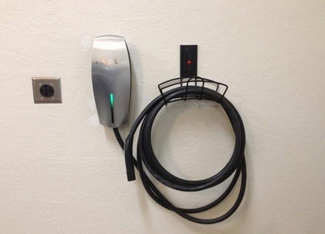 Wall-mounted EV charger with coiled black cable and outlet on a white wall.