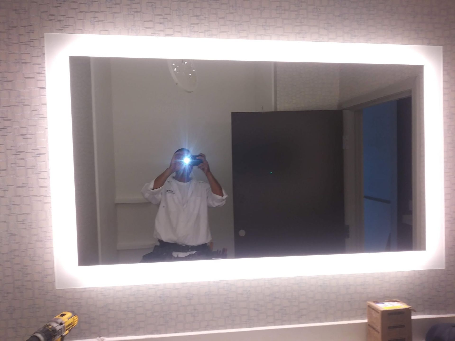 Person taking a mirror selfie in a lit room, with a dark wardrobe and small boxes visible.