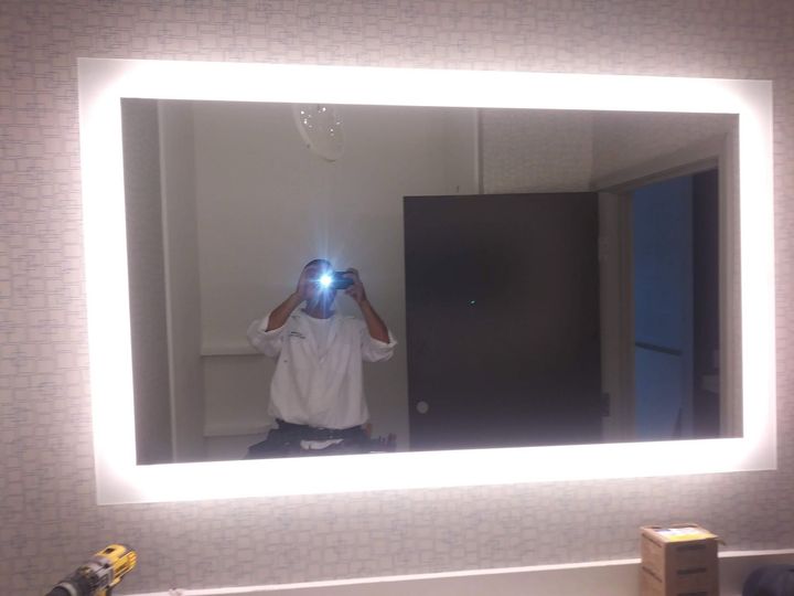 Person taking a mirror selfie in a bright bathroom with a glowing LED mirror and dark cabinet.