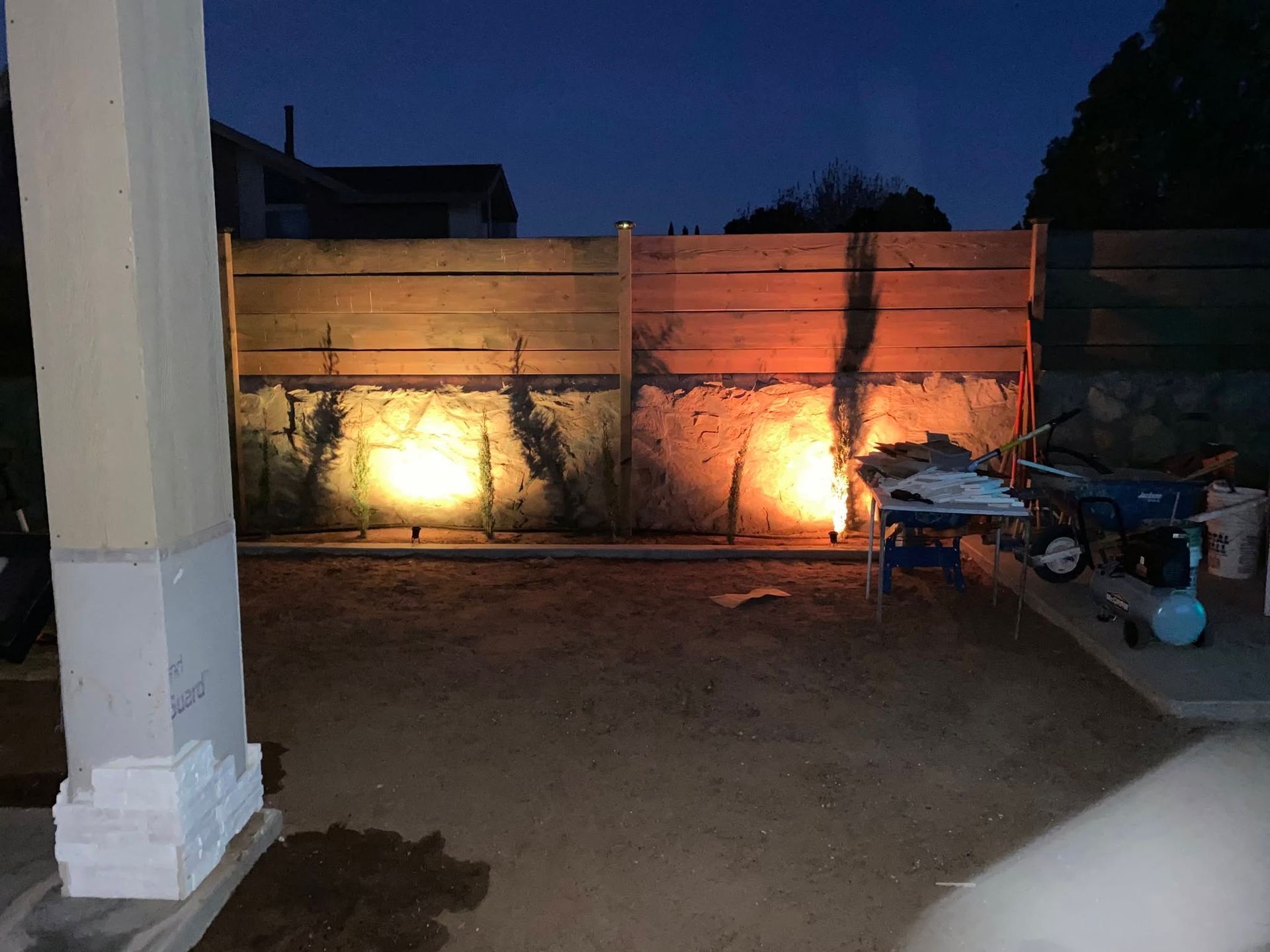 Nighttime outdoor patio with lit wall, white column, and a table with assorted items on the right