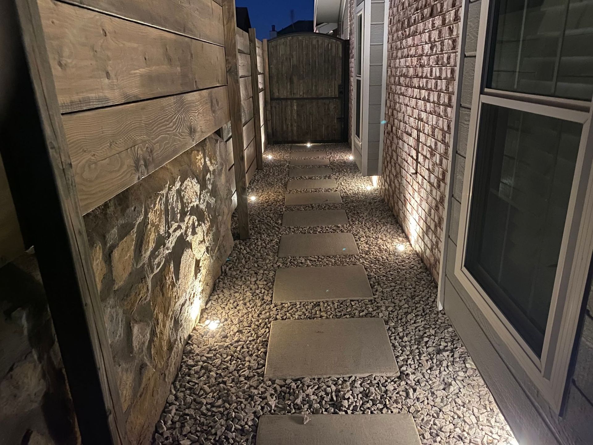 Narrow gravel side yard walkway between a wooden fence and brick house, lit by ground lights.