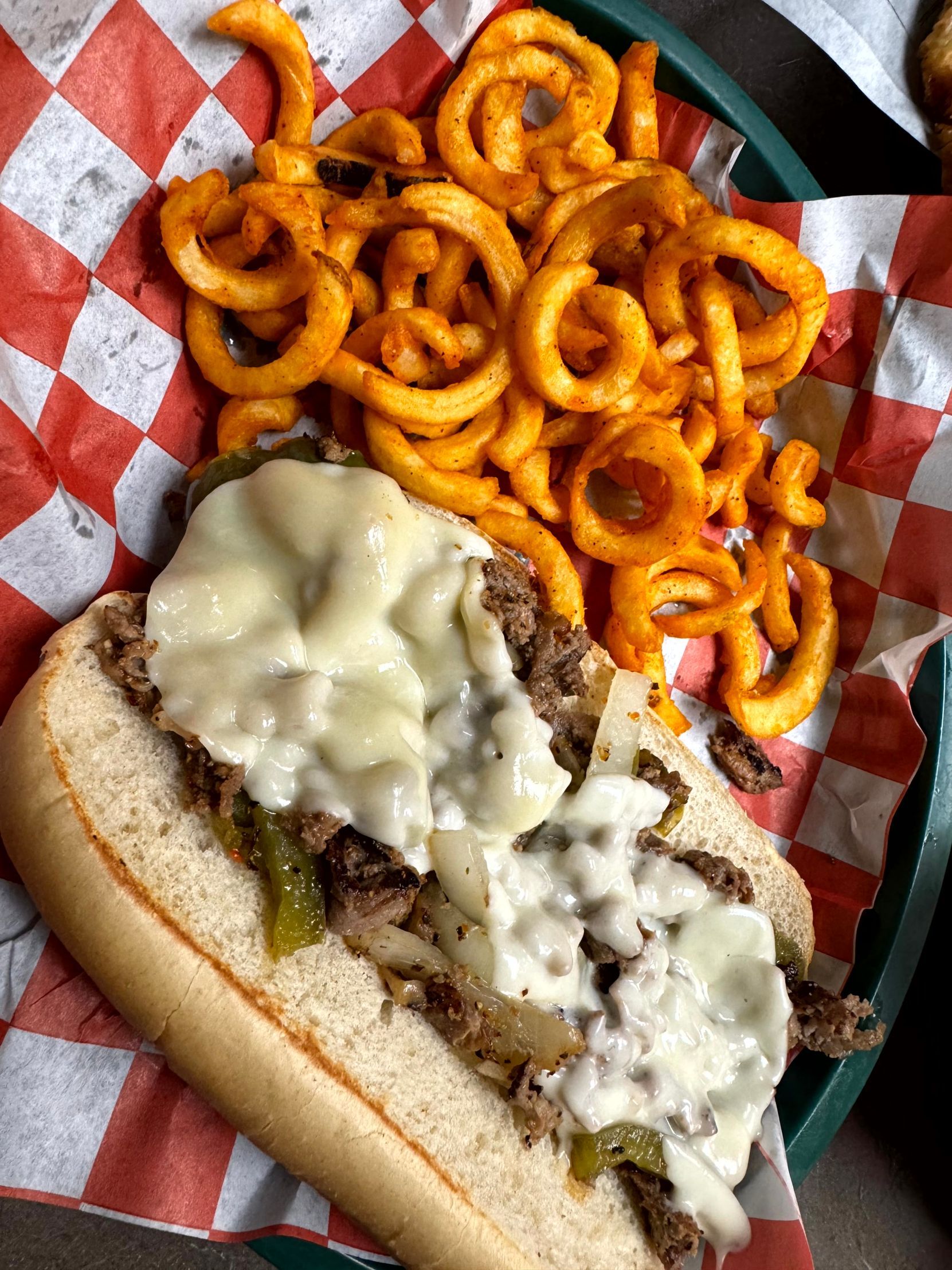 Philly cheesesteak sandwich with curly fries in a red-checkered basket.