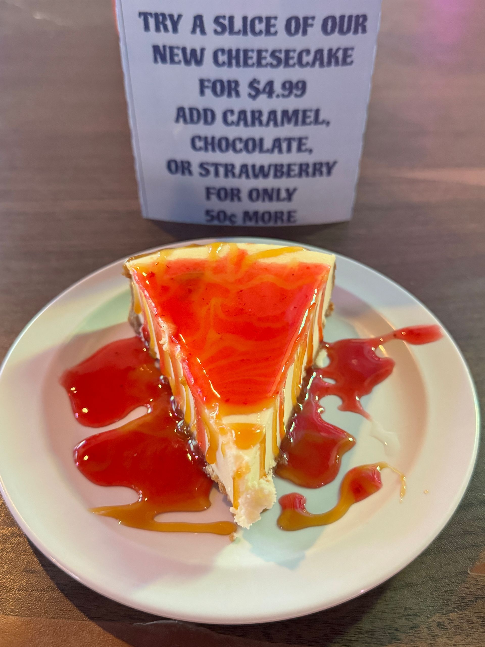 Slice of cheesecake with strawberry topping on a plate, with a sign advertising the dessert. Slice of cheesecake with strawberry topping on a plate, with a sign advertising the dessert.