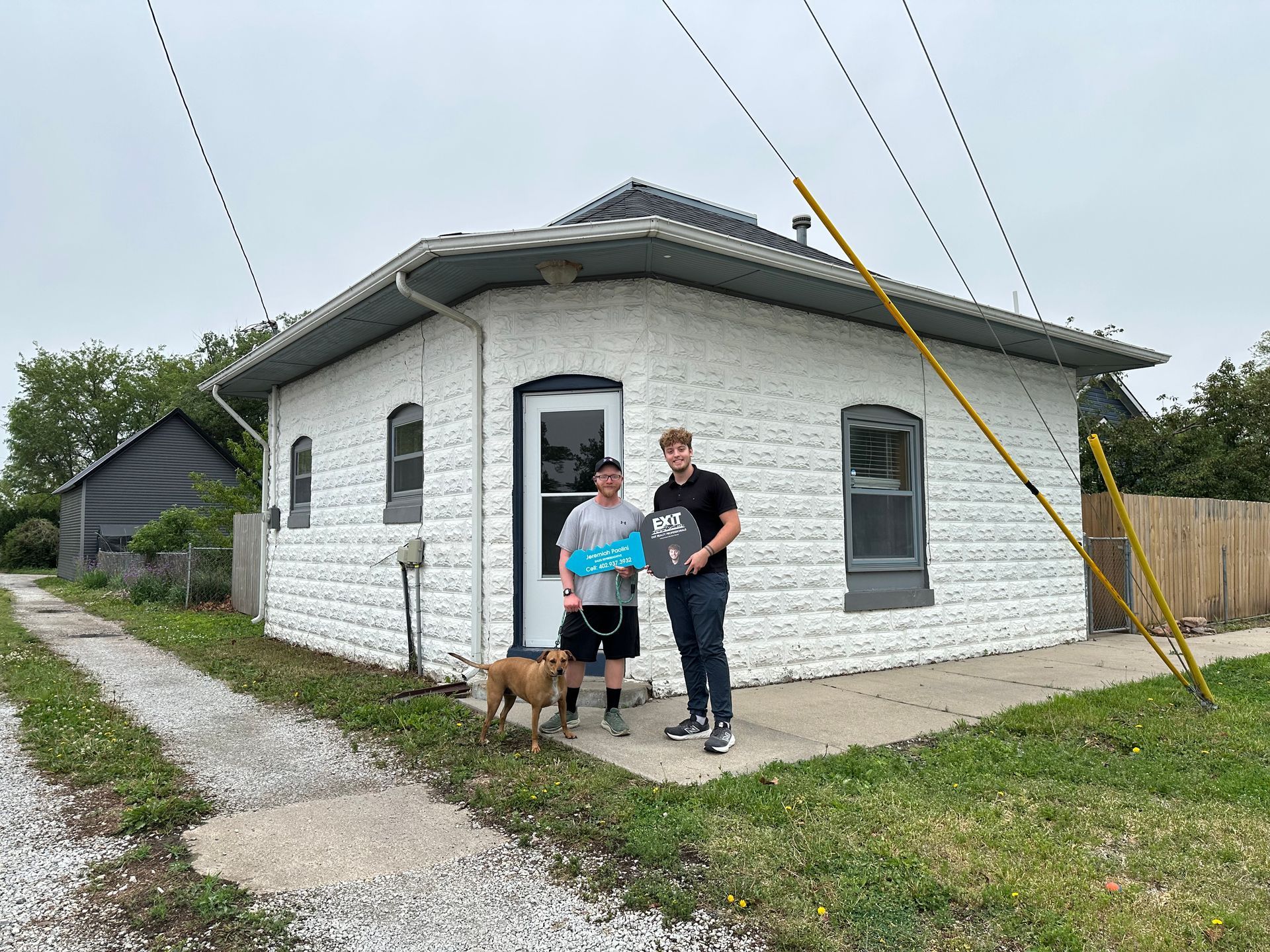 Two people and a dog standing beside a white house with a gravel path and grassy yard
