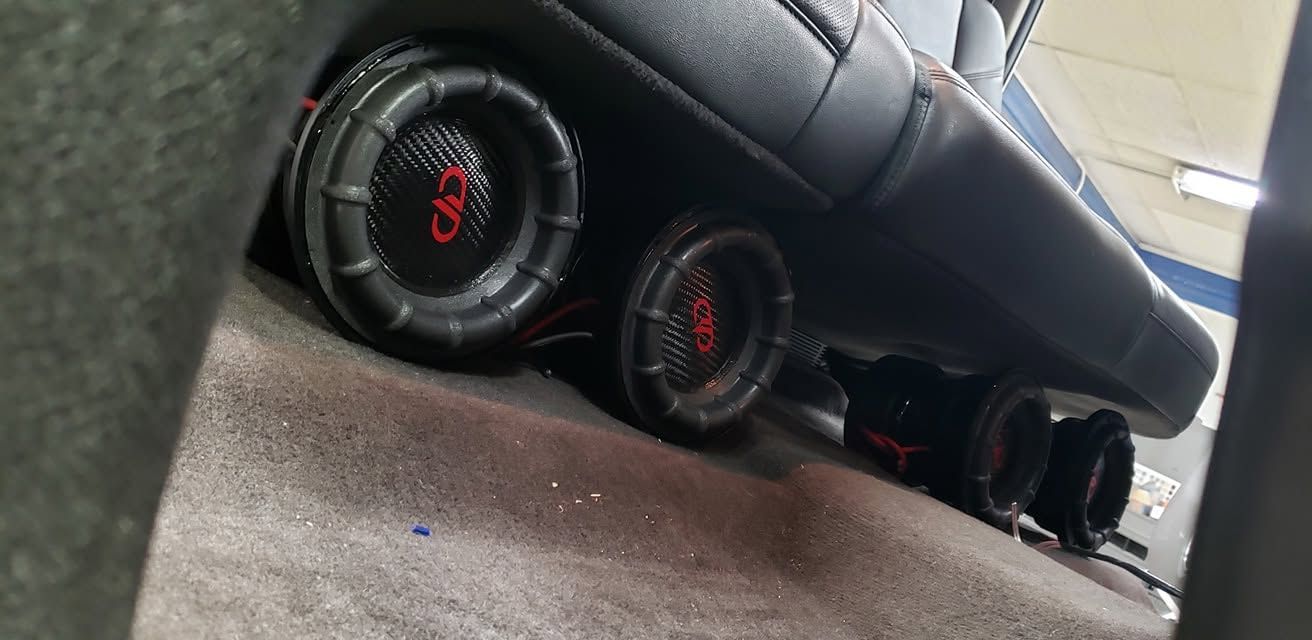Black subwoofer speakers lined up under a car dashboard on carpeted floor