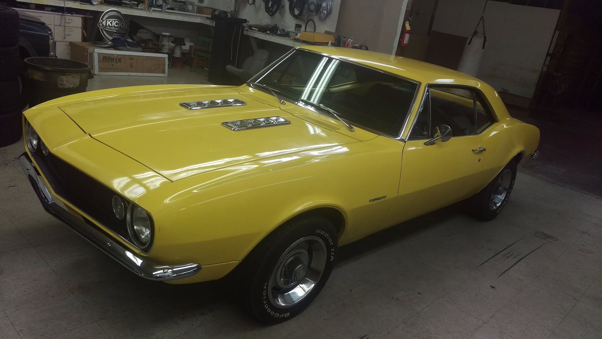 Yellow classic muscle car parked indoors, angled front-left view, with hood scoops and black racing stripes.