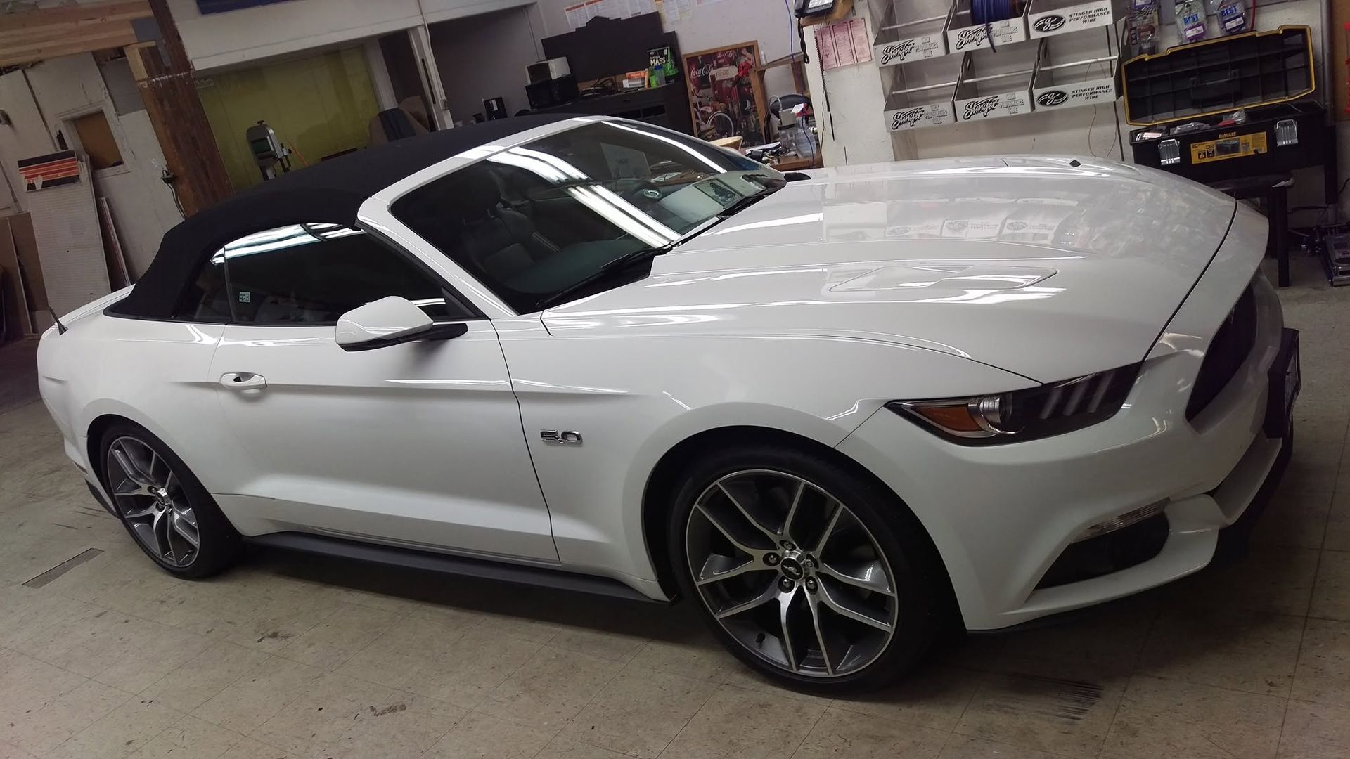 White Ford Mustang convertible parked indoors at a dealership or garage, shown in side view.