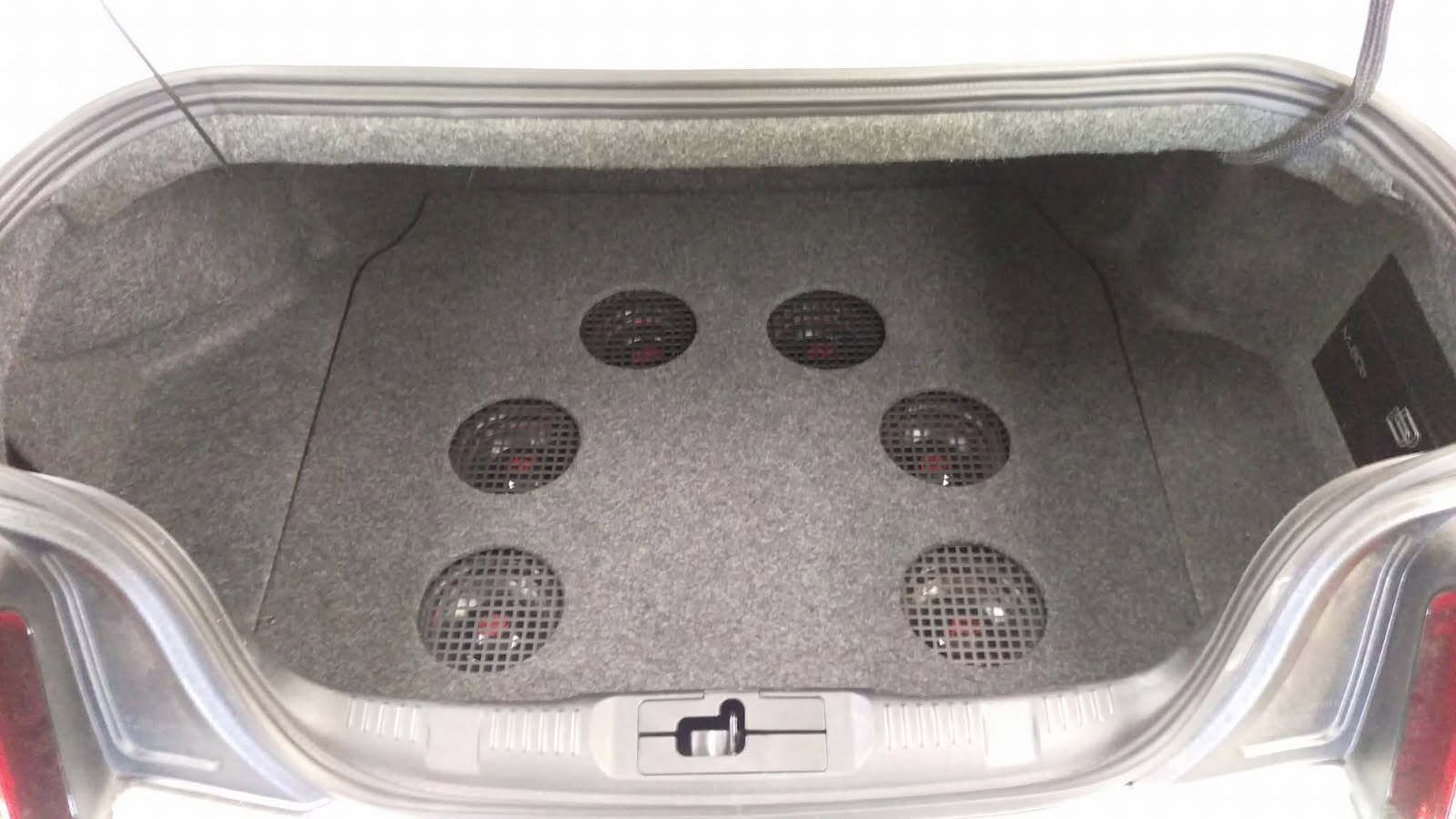 Car trunk lined with gray carpet and six round speaker grilles installed in the floor.