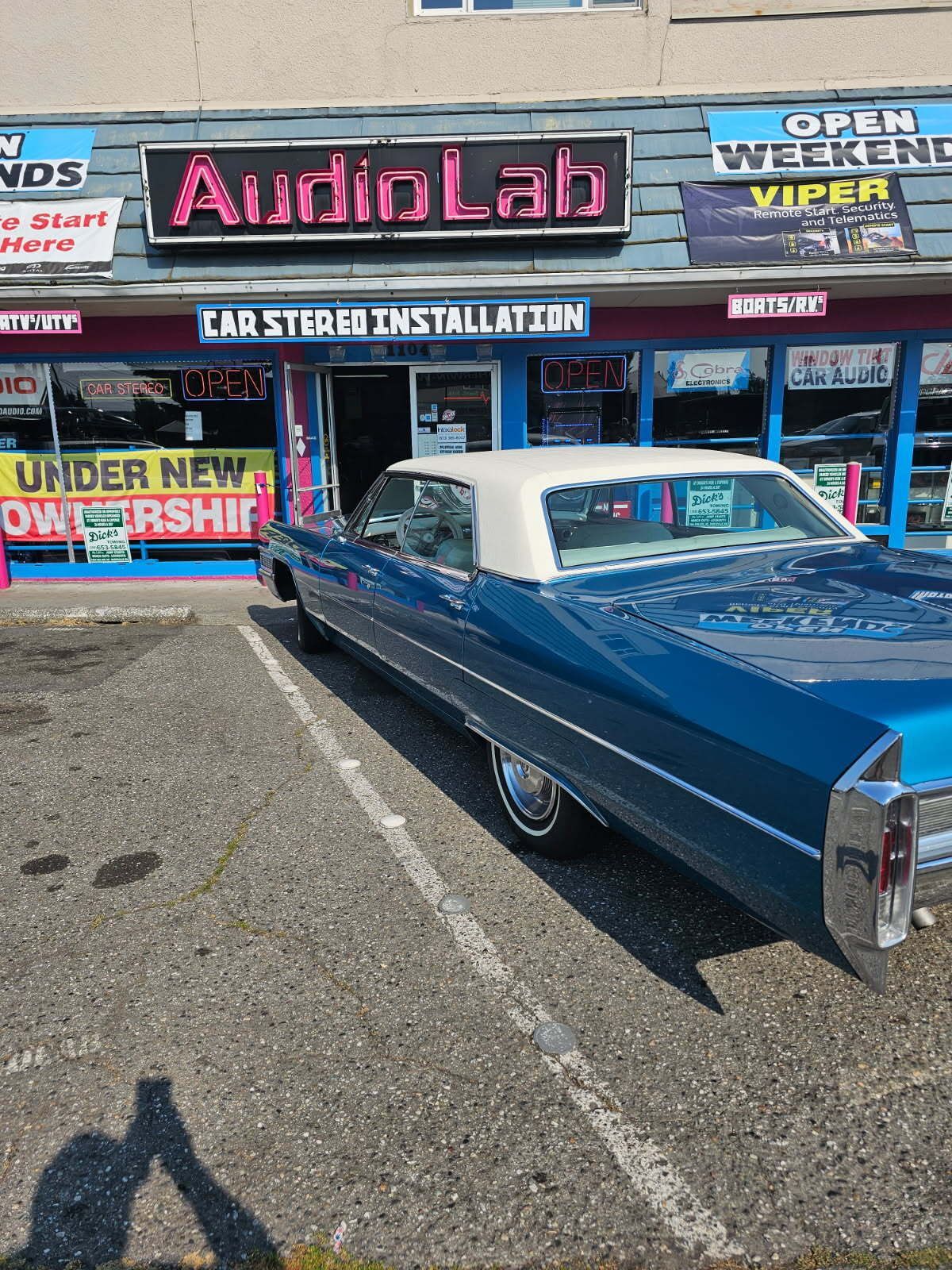 Blue classic car parked outside AudioLab auto stereo shop with neon sign and storefront windows