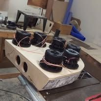Multiple black speakers mounted in a pale wooden box on a workbench, with wires and tools nearby