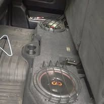 Car trunk with two large black subwoofers and a custom audio setup on a dark carpeted floor.