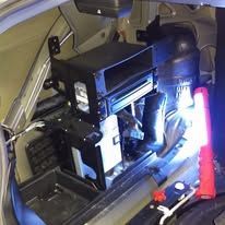 Equipment mounted inside a vehicle trunk, lit by a bright flashlight and red indicator light.