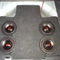 Car trunk subwoofer box with four black speakers on gray carpet.