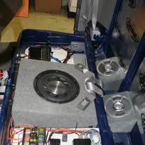 Car audio subwoofer installed in a blue-lined trunk with wiring and amplifier components nearby