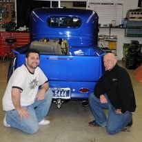 Two men kneel beside a bright blue vintage truck in a garage.