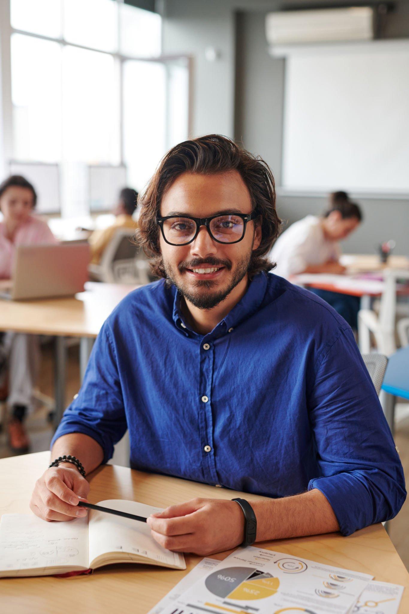 A man is sitting at a desk in a classroom reading a book.