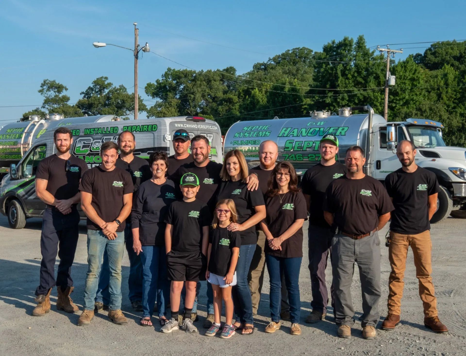 Group of people, including children, pose in front of two service trucks. All wear matching black shirts.
