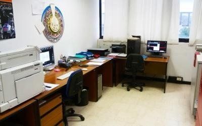 Ufficio con scrivanie, computer, stampanti e sedie lungo una parete con finestre e tende.