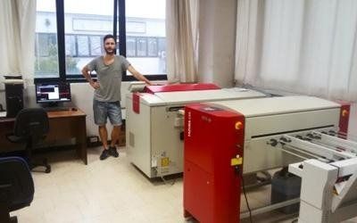 Persona in piedi in una tipografia accanto a una grande macchina industriale e a una scrivania vicino alle finestre.