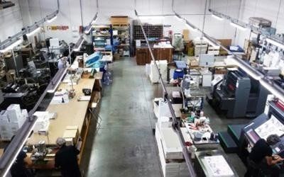 Officina industriale con lunghi tavoli da lavoro, macchinari e persone al lavoro in un luminoso spazio di fabbrica.