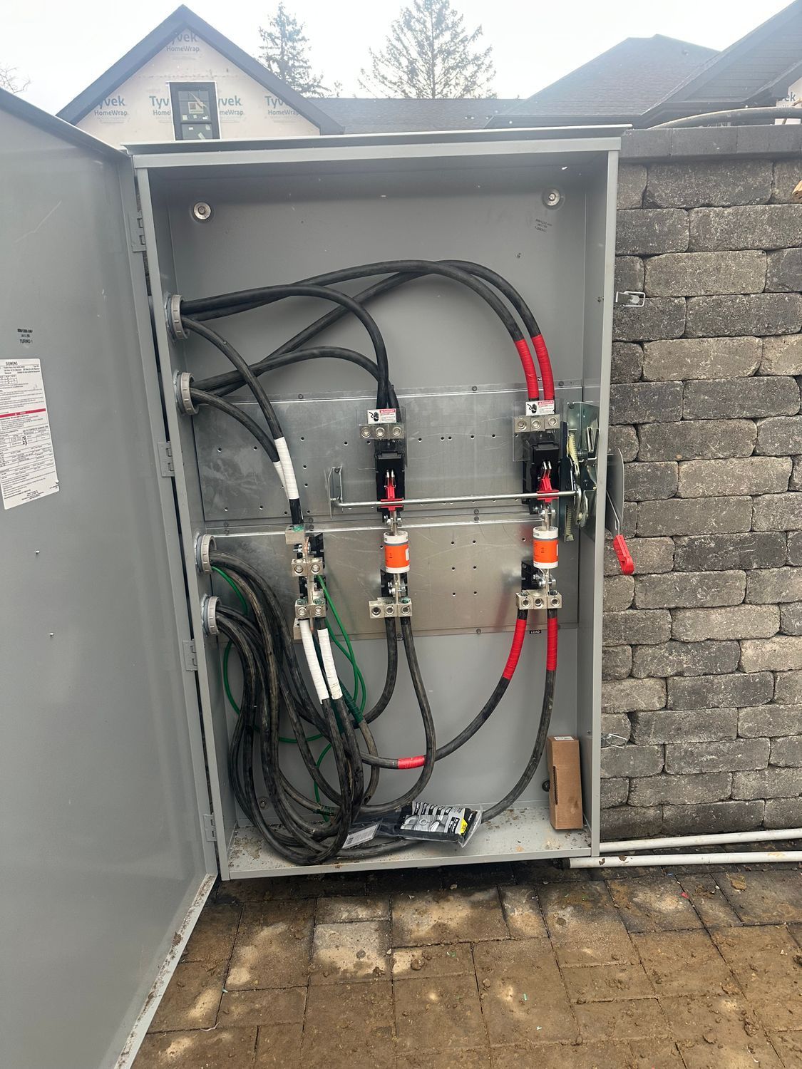 Open electrical panel with black, red, green wires and fuses. Mounted to brick wall, outside.