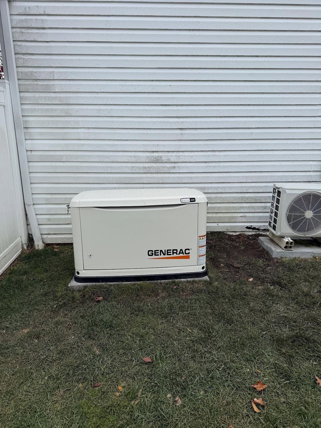 A Generac home standby generator sits on a concrete pad near an air conditioning unit and a white siding wall.