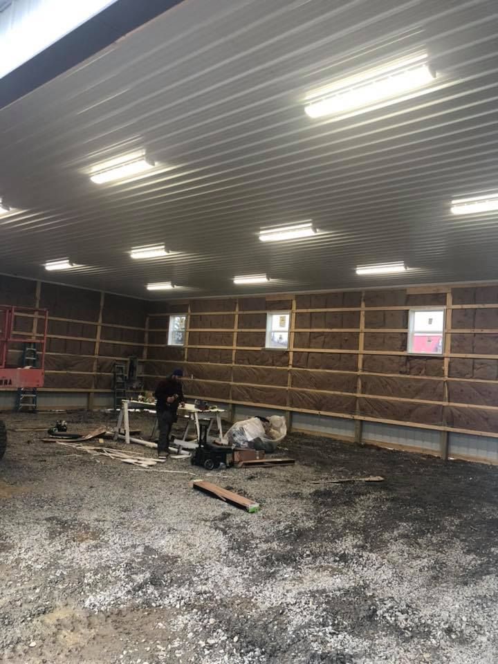 Interior view of a barn under construction, insulation installed on walls, exposed wood framing, fluorescent lights.
