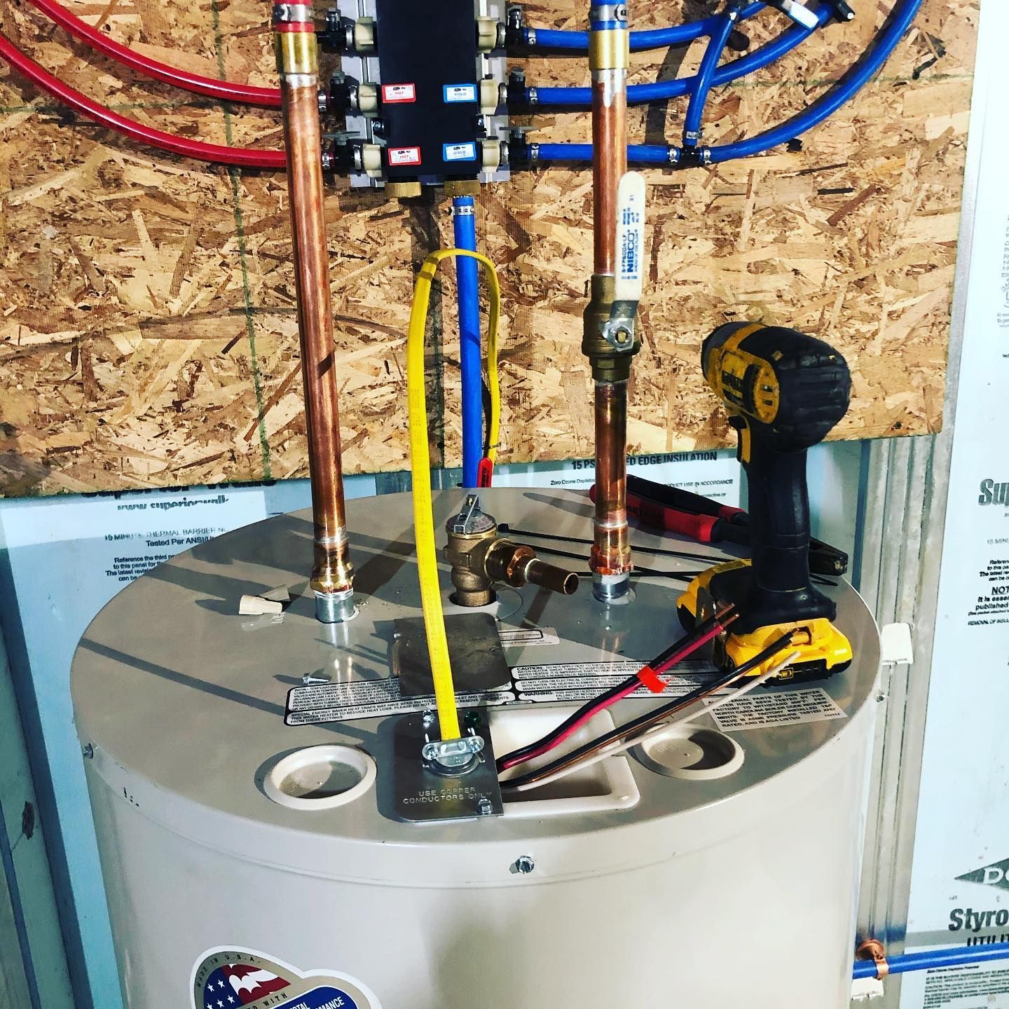 Water heater with copper pipes, a blue and red manifold, and a yellow electrical cable. A drill is visible.